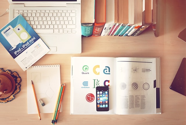 Image of books, stationery, mobile phone, and a laptop spread across a wooden table.
